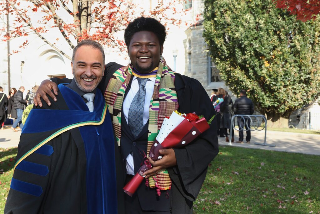 Professor Hossam Hassanein (left) posing next to Nana Boateng (right), both smiling and sharing a semi-embrace. They are posing in front of Grant Hall, surrounded by autumn trees shedding bright-orange leaves. Both are wearing graduation gowns. 
