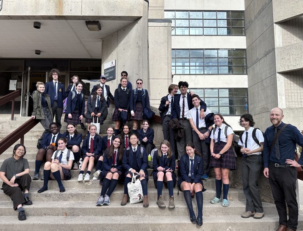 A group of High School Seniors (about 20-30 people) are posing on the steps of Goodwin Hall on a sunny afternoon, all wearing their school uniform. Two teachers, a woman and a man are also posing with the group, smiling. 