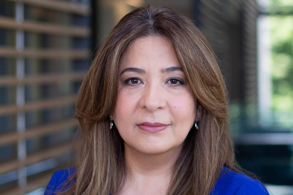 Professor Mousavi is posing for the camera in a portrait headshot. She is wearing a blue top and has her long brown hair down. The backdrop is a blurred interior of a winter garden in one of the buildings on Queen's campus
