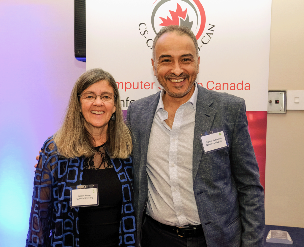 Professor Wendy Powley (left) and Queen's School of Computing Director Hossam Hassanein (right).
Professor Powley and Prof. Hassanein are standing close together posing for the camera with the CAN-CWiC conference banner backdrop. Both are wearing conference tags and smiling.